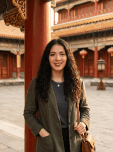 Mujer con cabello rizado y chaqueta verde oliva sonriendo frente a un patio del Palacio Imperial en Beijing, con arquitectura tradicional roja y dorada y faroles al fondo, luz cálida de atardecer.